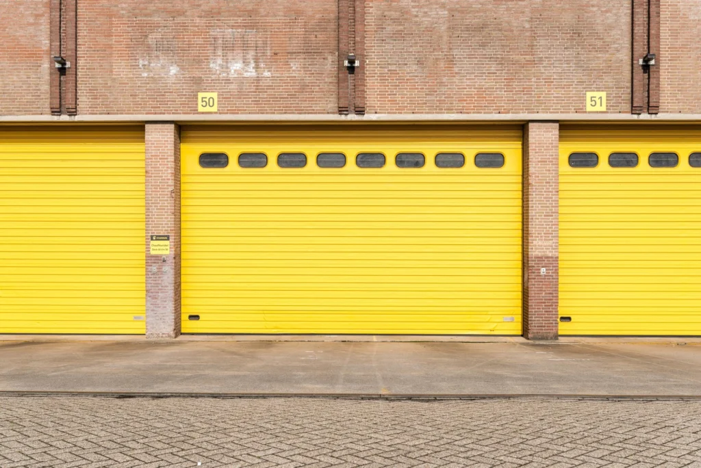 Two garage door technicians standing beside a residential garage, holding tools and smiling at the camera.